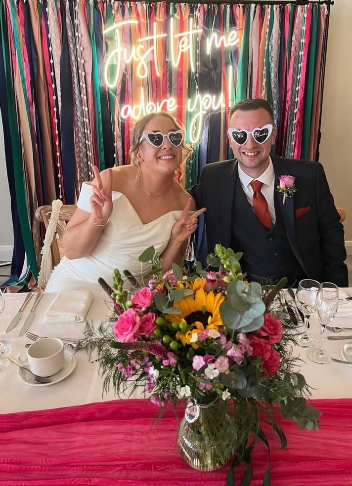 A happy bride and groom posing wearing sunglasses in front of a pink and green coloured ribbon backdrop with a neon sign attached which says ‘Just let me adore you.’