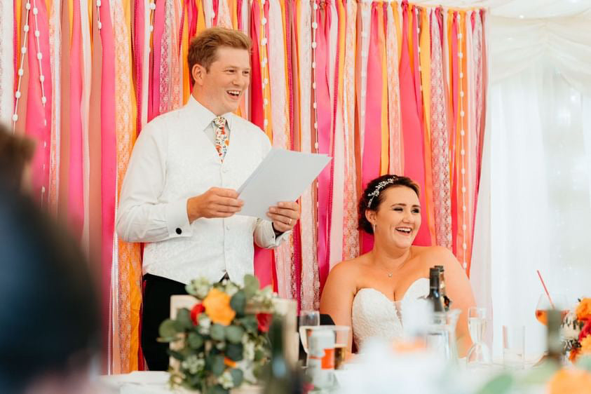 Colourful ribbon backdrop in pinks, oranges and white shown behind a head table with bride and groom seated in front.