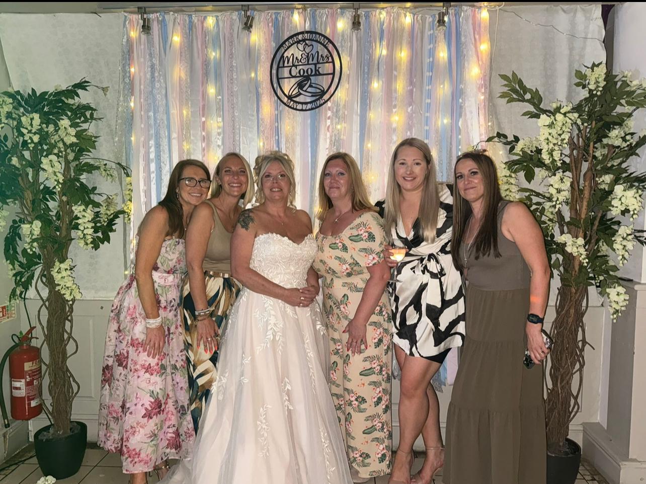 A group of ladies together with the bride posing in front of a ribbon backdrop which is white, pale pink and pale blue with fairy lights.
