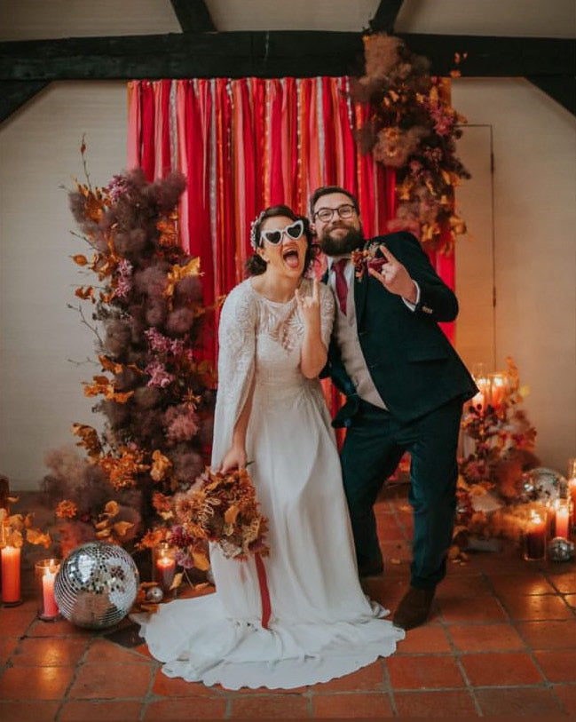 A bride and groom posing wearing sunglasses in front of a ribbon backdrop Im warm tones of pinks, oranges and reds. There is an arrangement of dried flowers at each side of the backdrop and pillar candles and disco balls at the base.