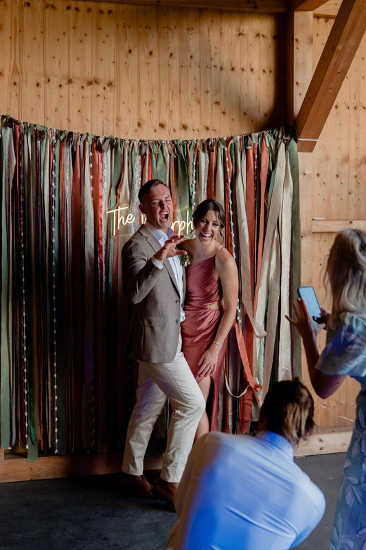 A backdrop displayed on a barn wall in copper and olive green colours. A couple are posing in front of the backdrop, the copper colour matches the woman’s bridesmaid dress.