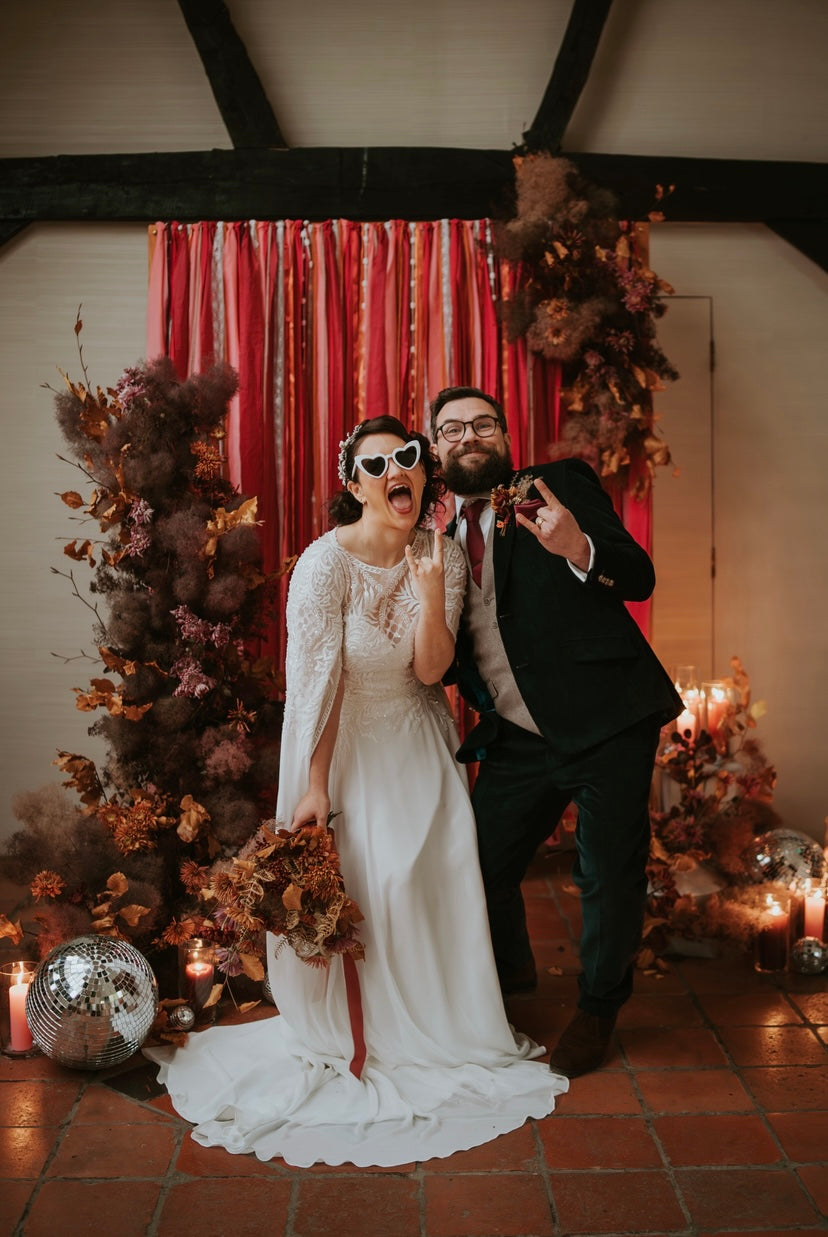 Bride and groom posting in front of a ribbon backdrop in autumnal shades. Also on the backdrop is a dried floral arrangement.