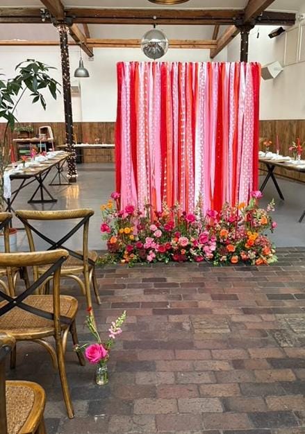 A colourful ribbon backdrop in reds and pinks with white lace and various textures. The backdrop is at the end of a wedding aisle and has a beautiful display of flowers at the base in matching colours. 