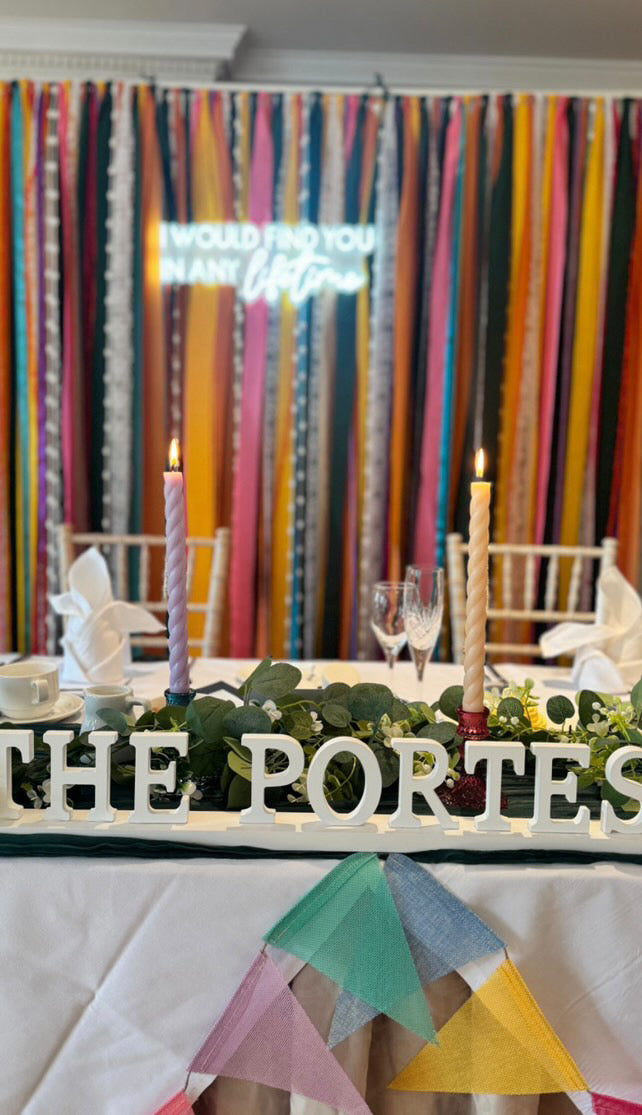 A colourful ribbon backdrop in vibrant shades of yellow, pink, green and blue set behind a wedding head table. Backdrop has a neon sign attached. 