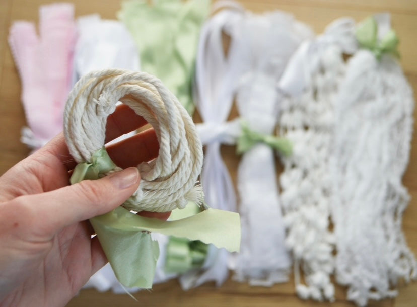 A DIY garland kit showing an assortment of ribbons and fabrics tied together in separate coloured bundles with a tied bundle of rope in the foreground. 
