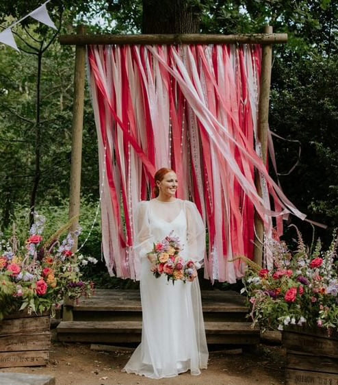 A ribbon backdrop on shades of pink attached to a large wooden frame, flowers each side and bride posing in front. 